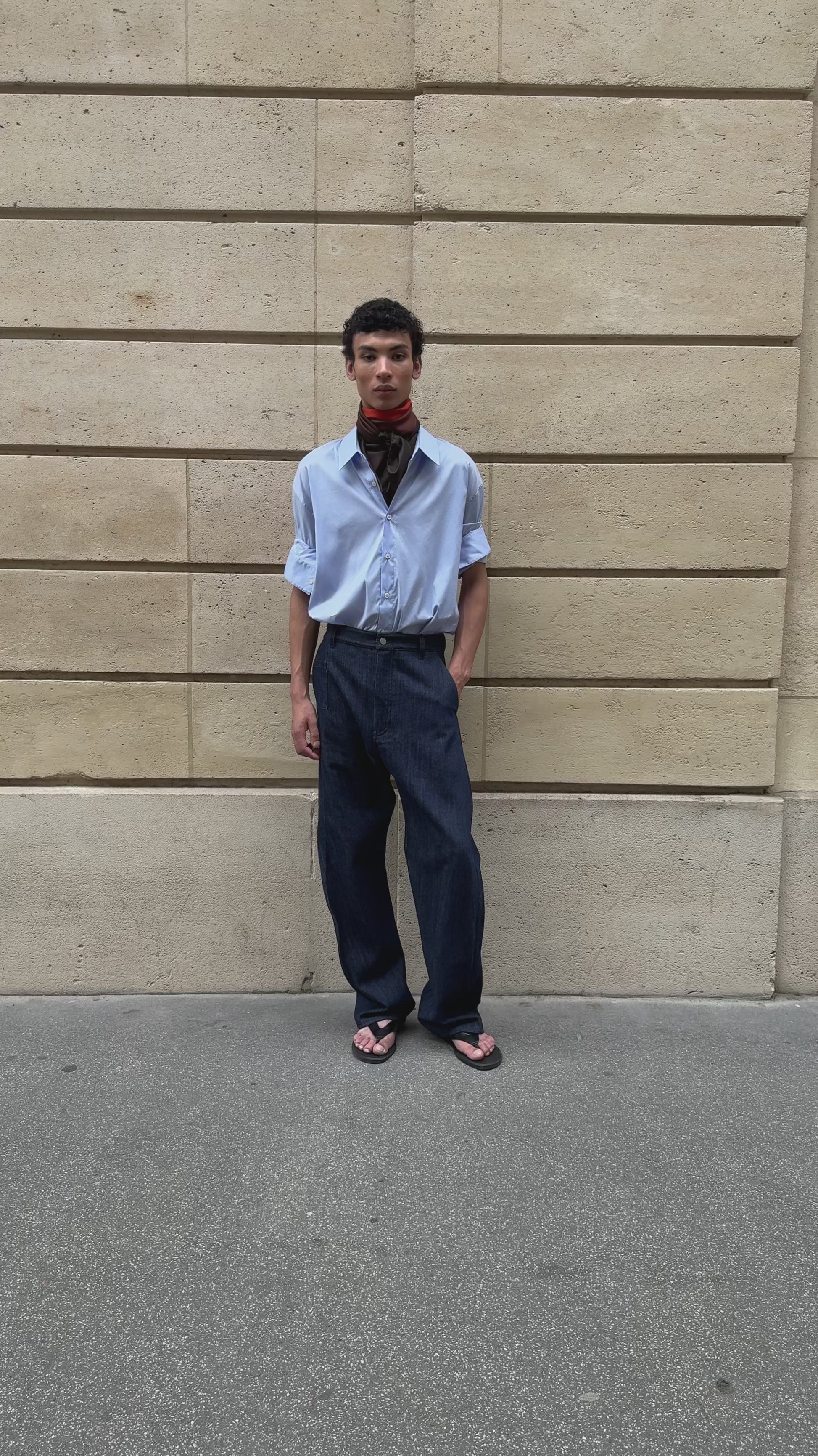 Male model in front of a stone wall wearing APAR Edition 002 - an oversized shirt, a carre 90 printed scarf and twisted leg trousers in denim